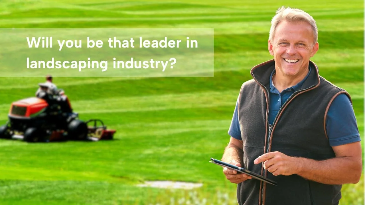 A smiling man holding a tablet on a striped lawn with a mower behind him